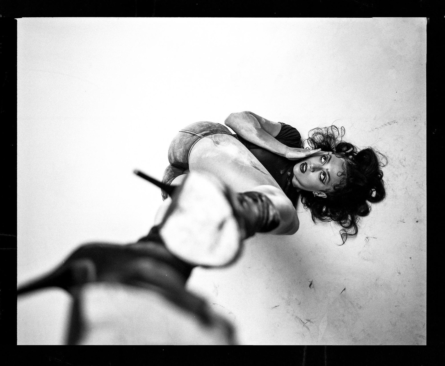 A woman with dark, curly hair and dark lipstick lies on a light surface, looking up at the viewer.