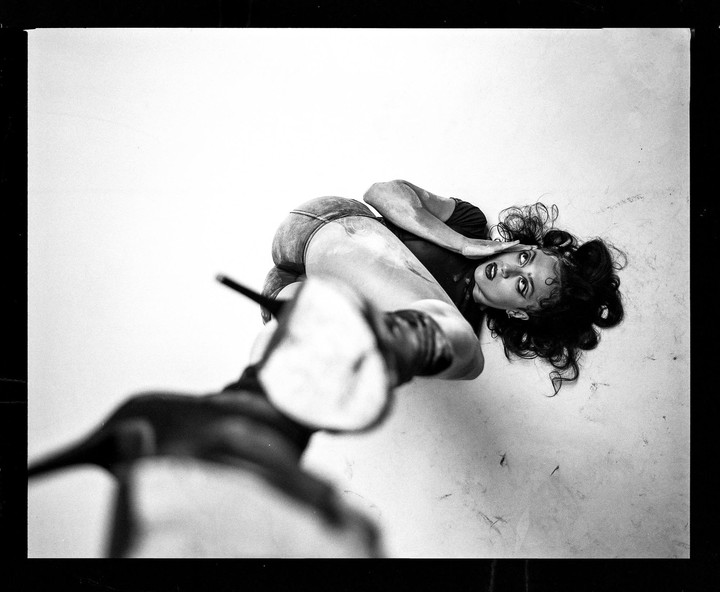 A woman with dark, curly hair and dark lipstick lies on a light surface, looking up at the viewer.