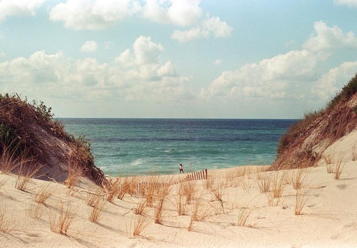 A woman walks along a sandy beach with dunes and the ocean under a cloudy sky.