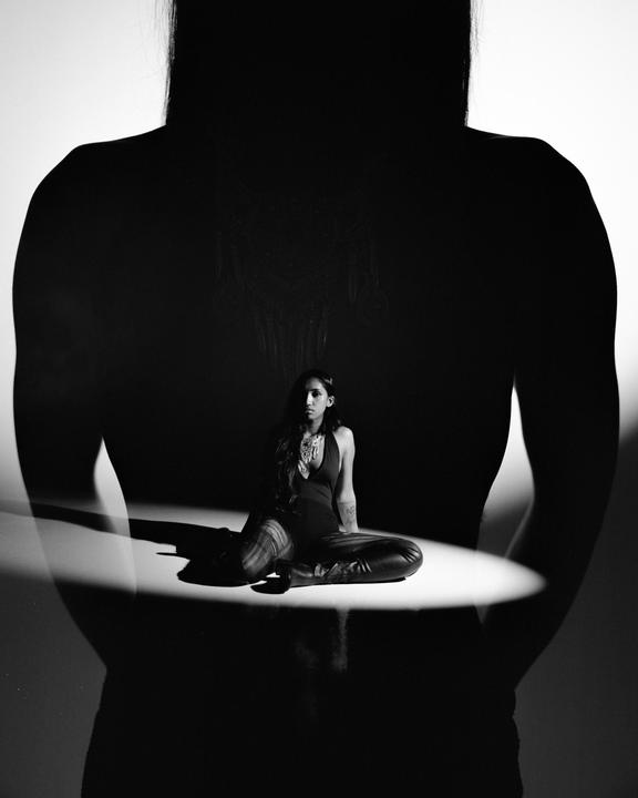 A woman sits on the floor in a spotlight, framed by a large silhouette of a person.