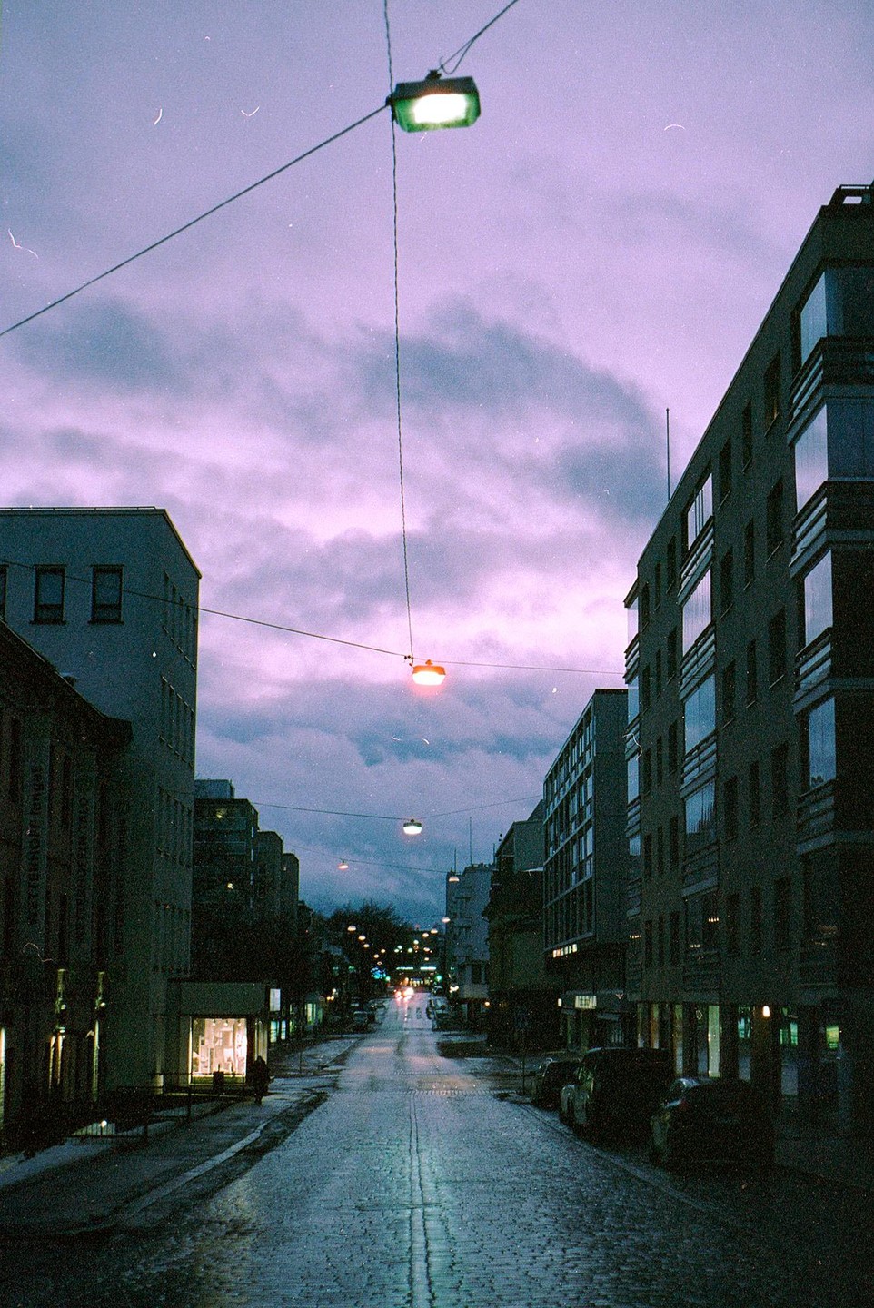 A street with buildings and cars is illuminated by streetlights under a purple sky at night.