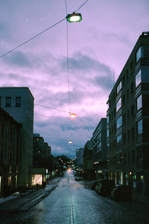 A street with buildings and cars is illuminated by streetlights under a purple sky at night.