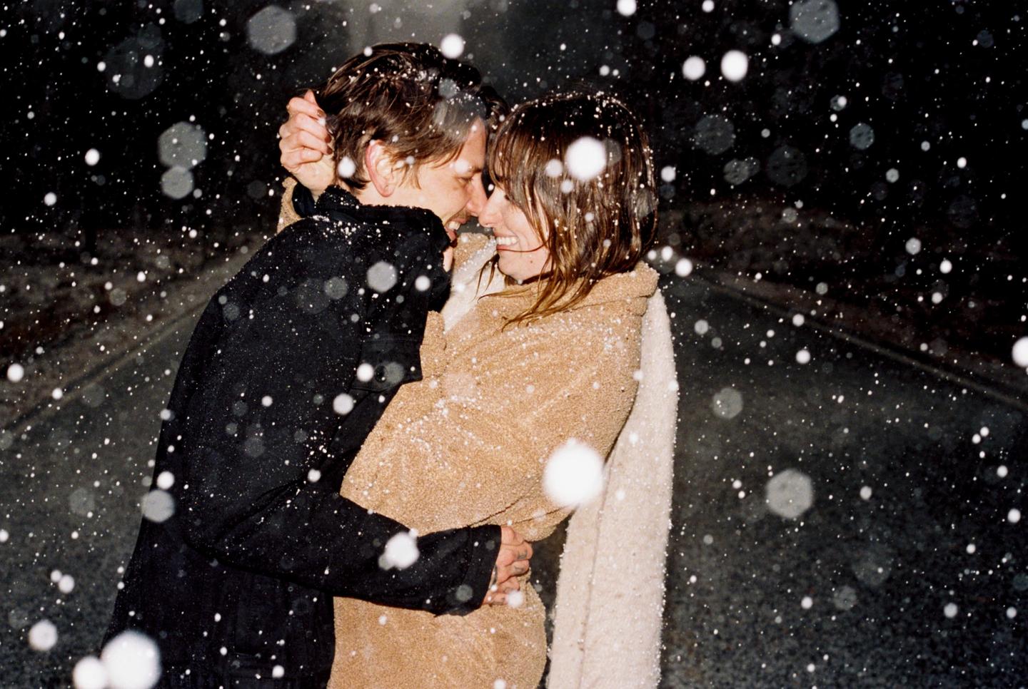 A couple embraces while snow falls around them at night.