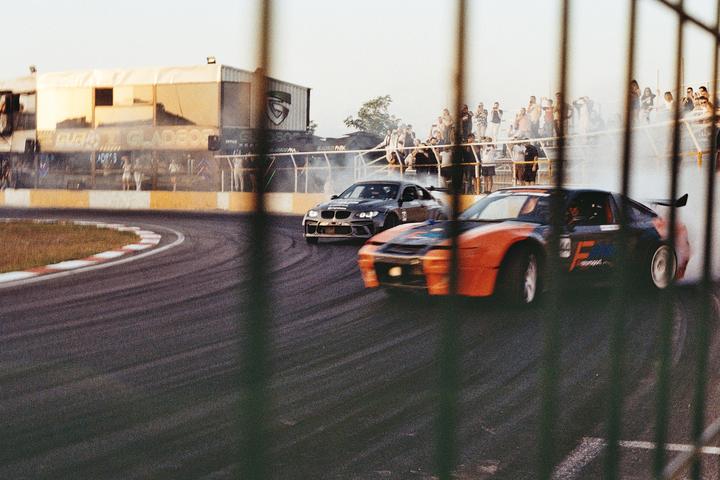 Two race cars drift around a track with spectators watching from behind a fence.