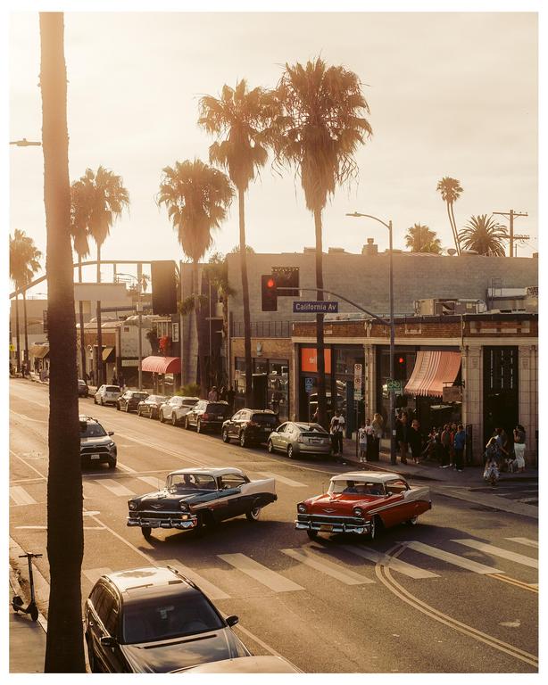 Two vintage cars drive on a street lined with palm trees and buildings under a hazy sky.