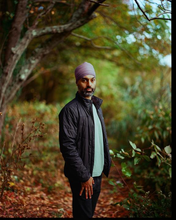 A man wearing a turban and a jacket stands in a forest with autumn foliage.