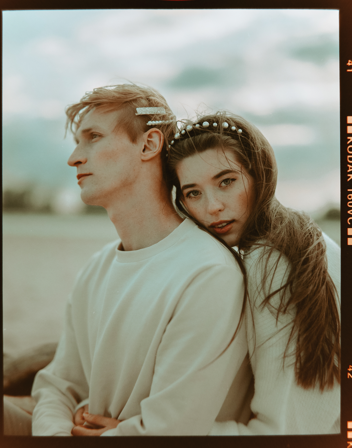 A woman rests her head on a man's shoulder, both wearing light-colored sweaters, with the woman looking towards the viewer.