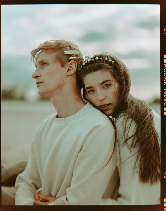 A woman rests her head on a man's shoulder, both wearing light-colored sweaters, with the woman looking towards the viewer.