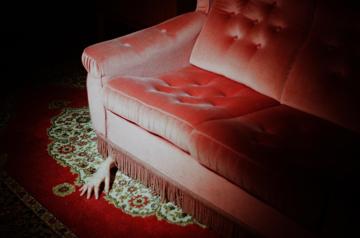 A hand reaches out from under a pink sofa with fringe onto a red patterned rug.