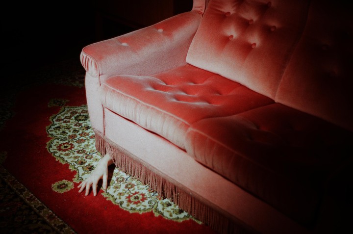 A hand reaches out from under a pink sofa with fringe onto a red patterned rug.