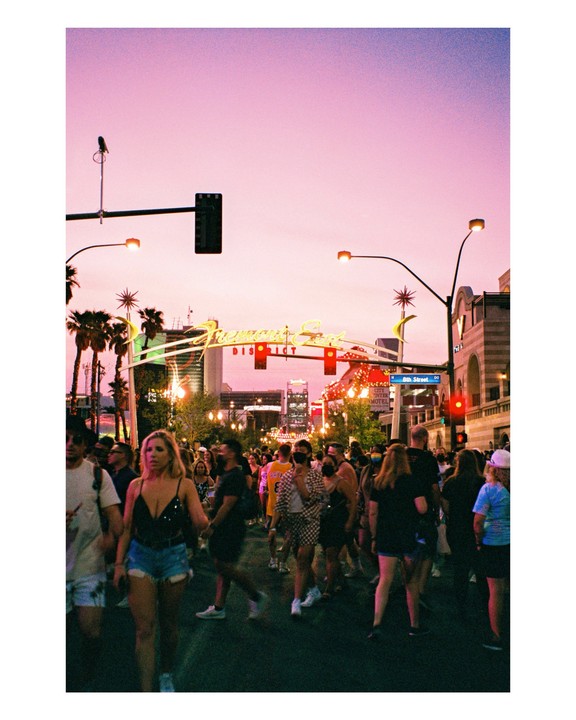 A crowd of people walk down a street at dusk with neon signs and palm trees visible.