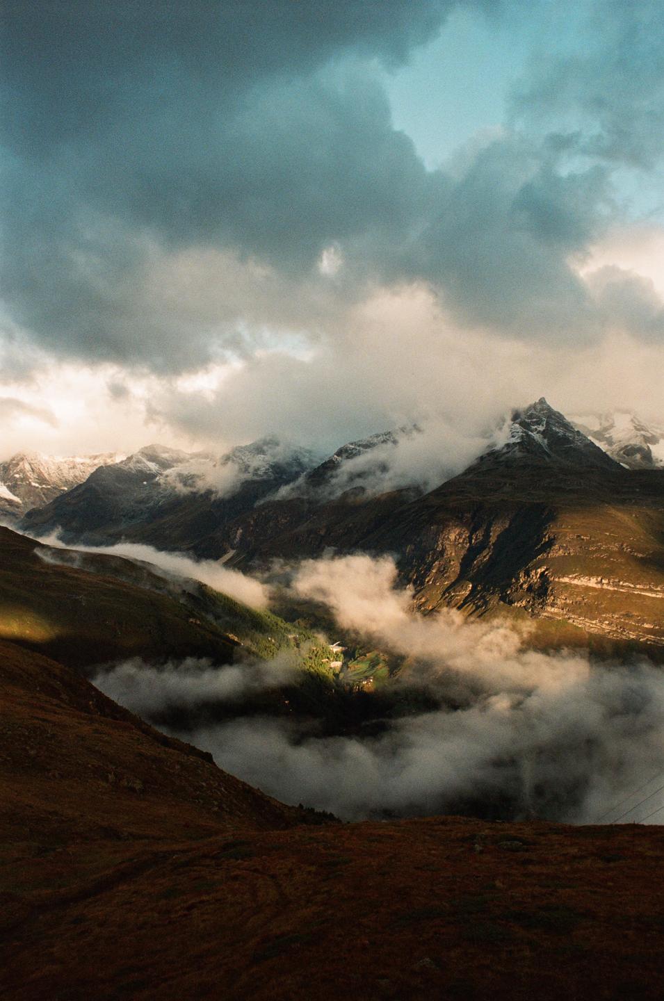Clouds and fog cover a valley and mountains with patches of snow.