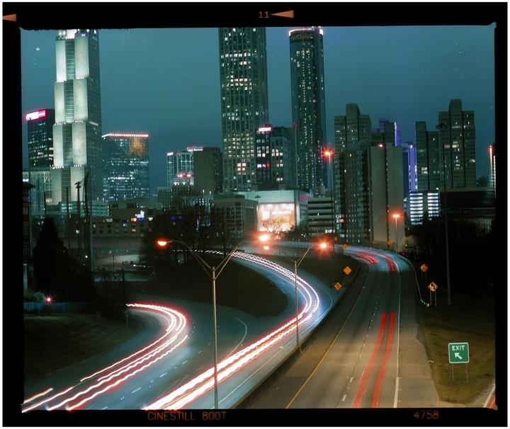 A long exposure captures light trails from vehicles on a highway at night, with a city skyline in the background.
