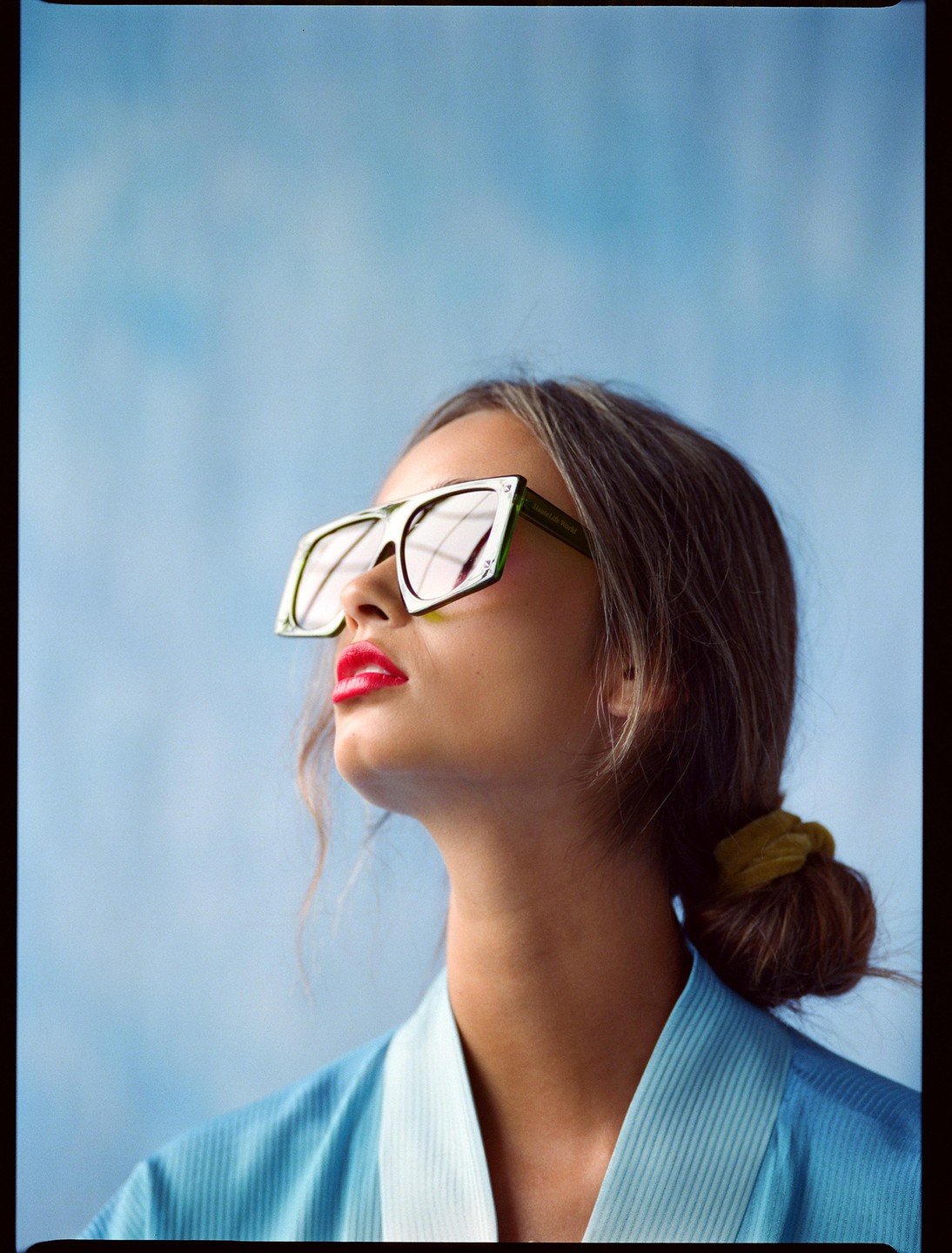 A woman with red lipstick and green sunglasses looks up against a blue background.