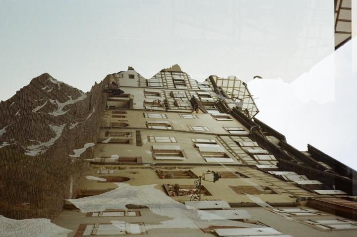 A double exposure shows a tall building with many windows and a cobblestone mountain range under a bright sky.