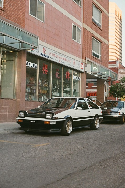 Two white and black cars are parked on a street in front of a building with a store on the ground floor.