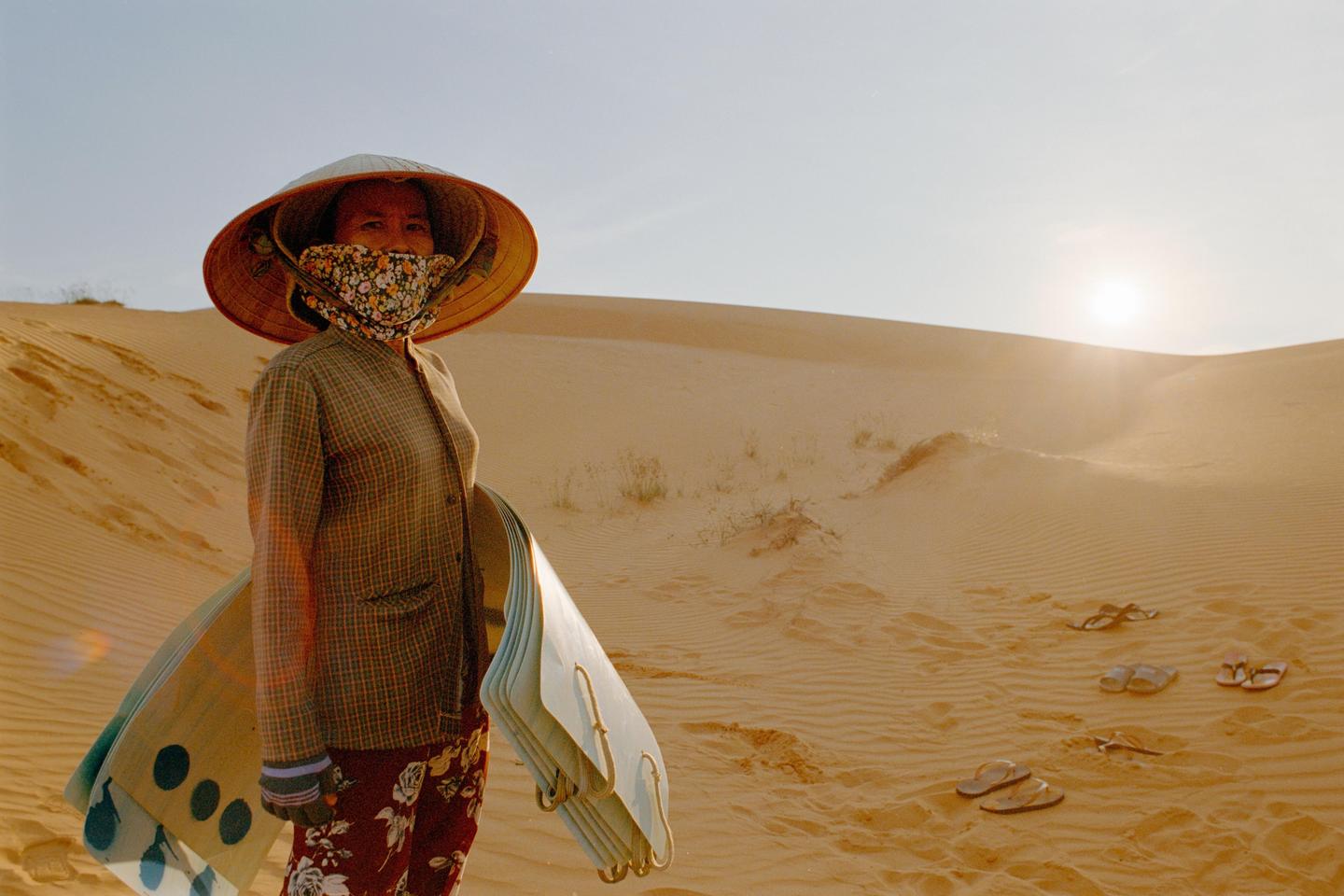 A woman wearing a conical hat and a floral face covering stands in a desert landscape with sand dunes and a bright sun.