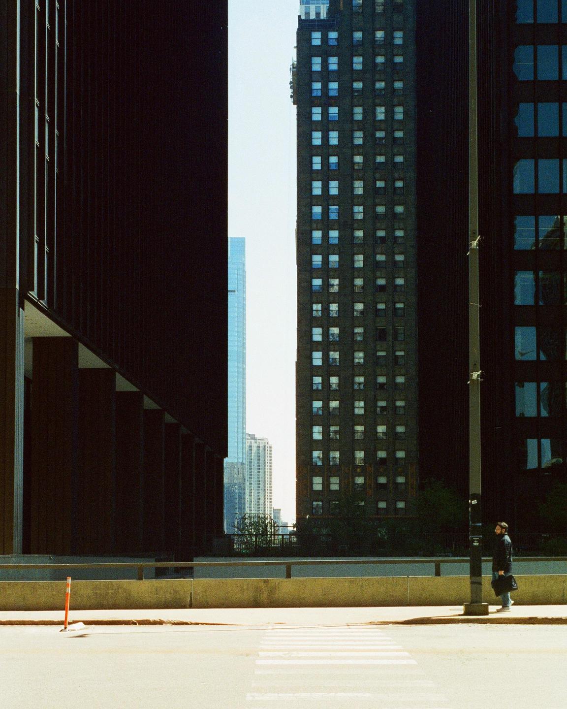 A man stands on a street corner next to a lamppost, with tall buildings and a crosswalk visible.