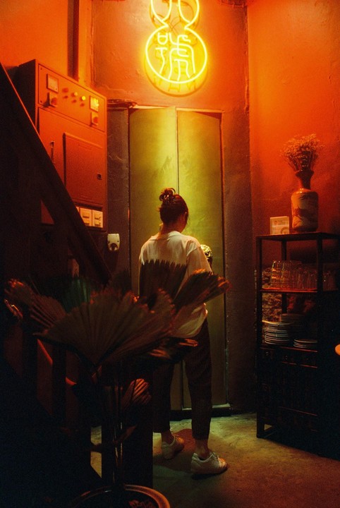 A woman stands in a dimly lit room with red and green walls, illuminated by a neon sign.
