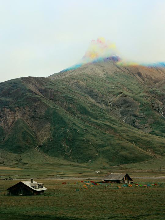 A colorful cloud hovers over a mountain with two houses and a herd of cattle in the foreground.