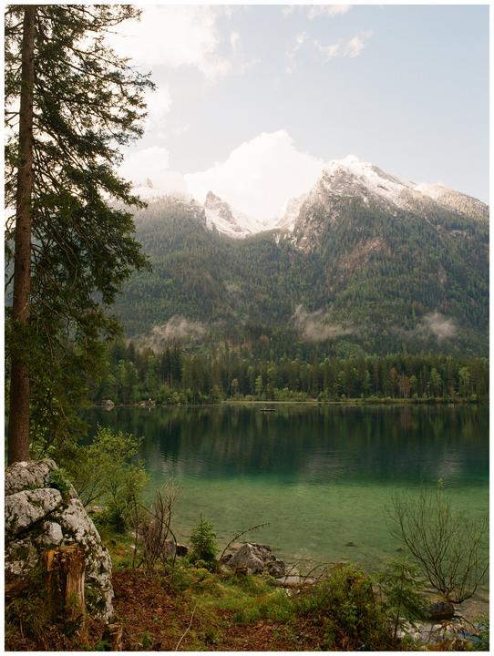 A clear lake reflects a dense forest and snow-capped mountains under a cloudy sky.