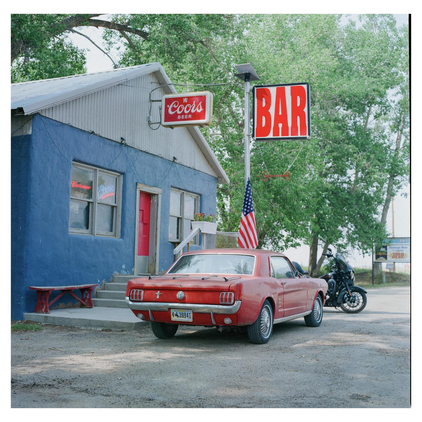 A red car is parked in front of a blue building with a "BAR" sign and an American flag.