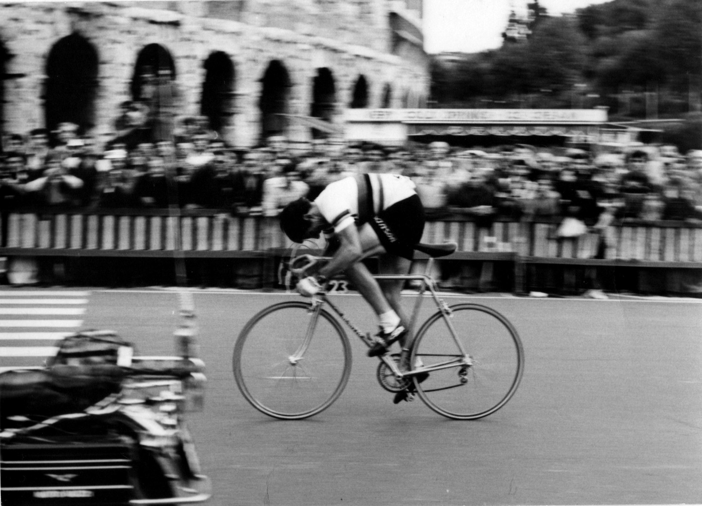 A man on a racing bicycle speeds past a crowd of spectators and an ancient Roman structure.