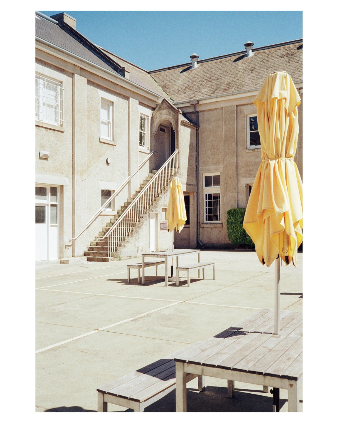 A courtyard with tables, benches, and yellow umbrellas is surrounded by a building with a staircase.