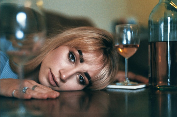 A woman with blonde hair and bangs rests her head on a reflective surface next to a glass of wine and a bottle.