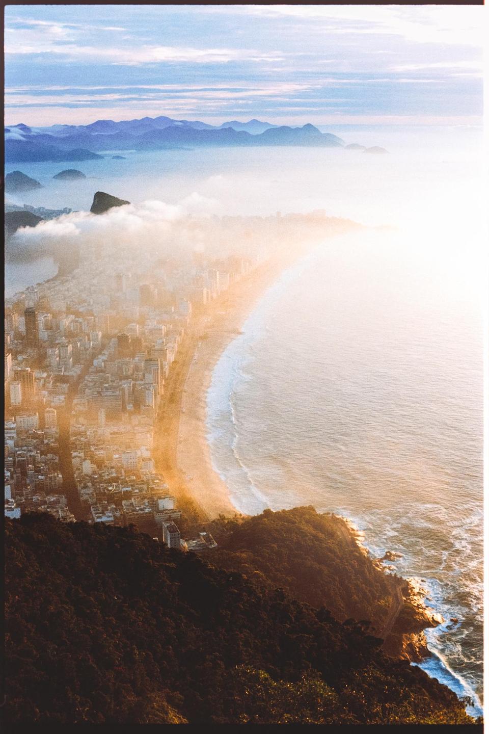 A city and beach are visible from a high vantage point, with mountains and clouds in the distance.
