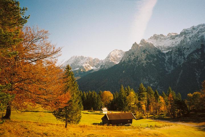 A small wooden cabin sits in a golden field with autumn trees and snow-capped mountains in the background.