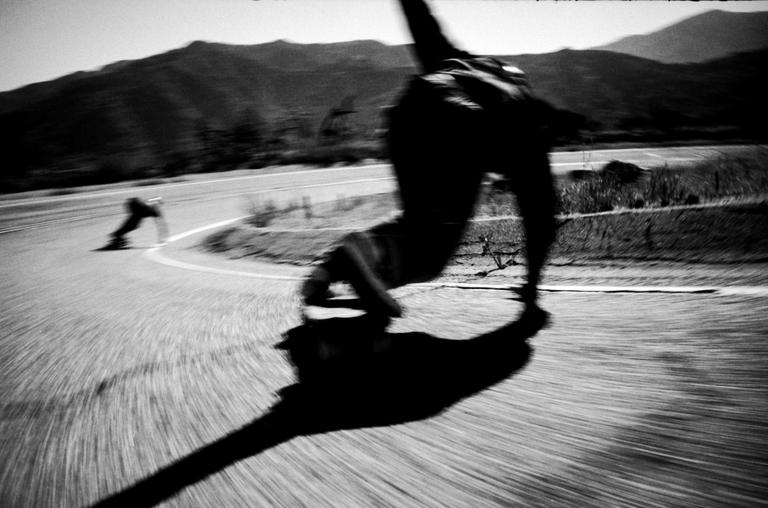 A person on a skateboard leans into a turn on a mountain road, casting a long shadow.