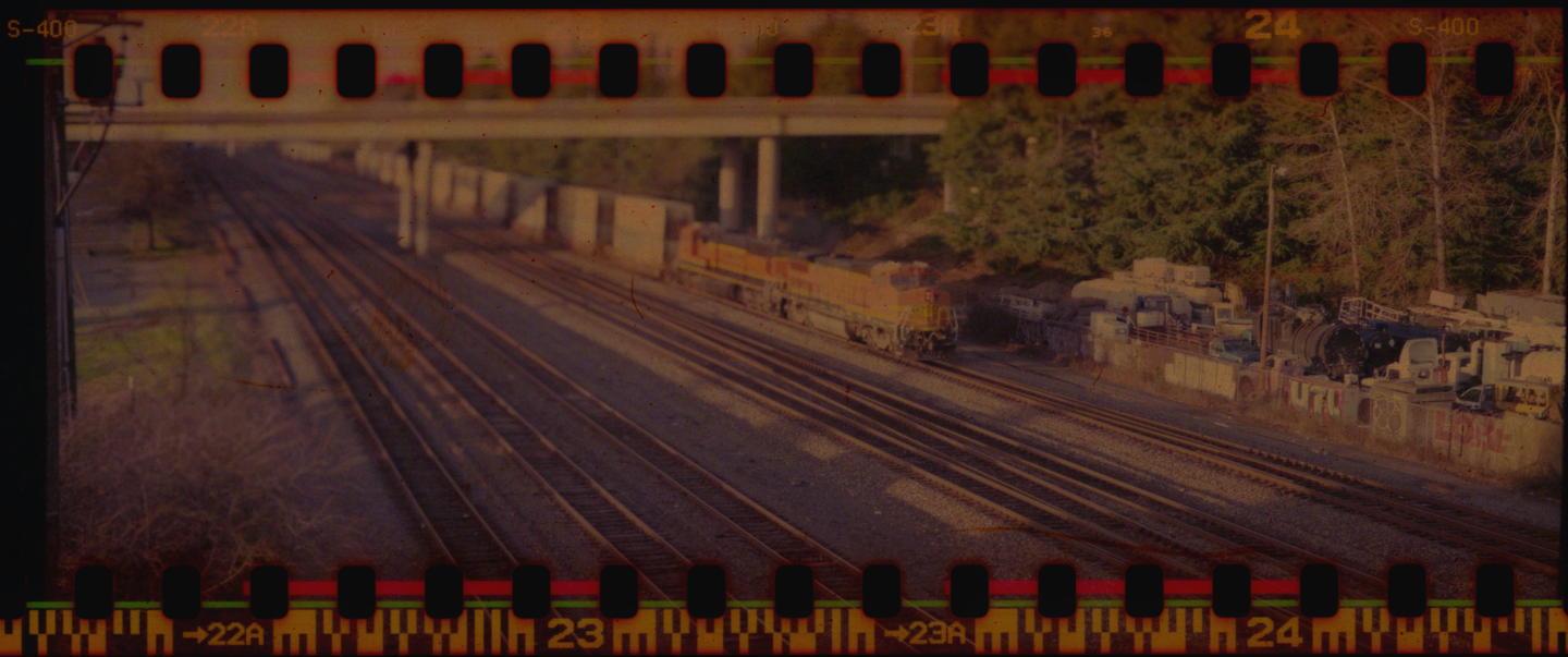 A train is visible on multiple tracks next to a wall covered in graffiti, with trees and an overpass in the background.