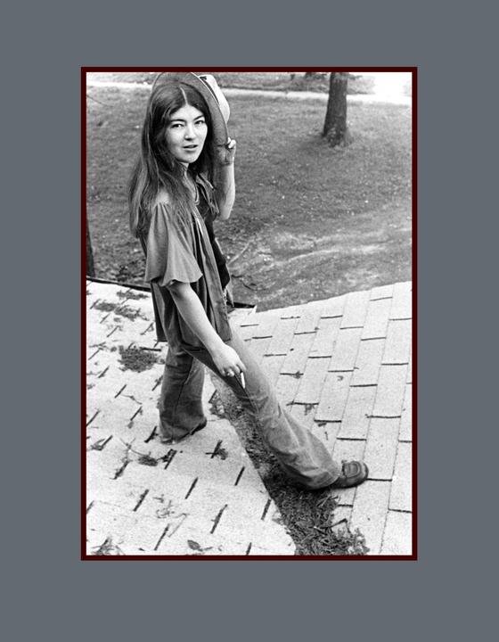 A woman stands on a shingled roof, holding a hat and looking over her shoulder.
