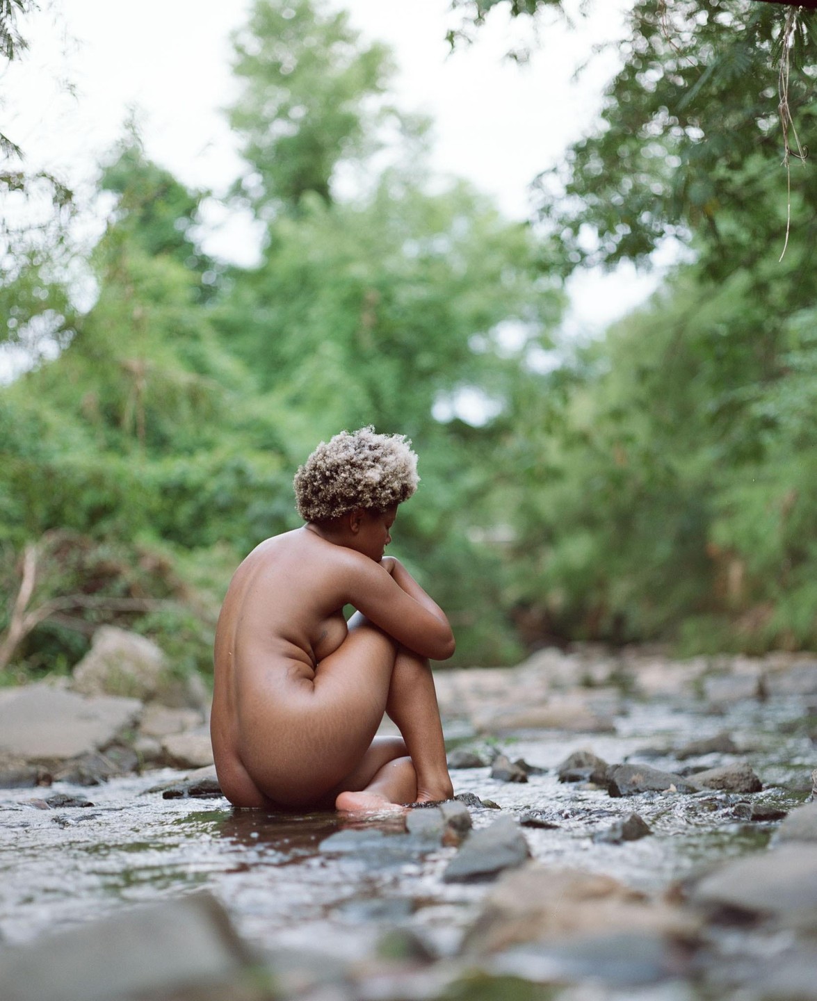 A nude woman with curly hair sits in a shallow rocky stream with green foliage in the background.