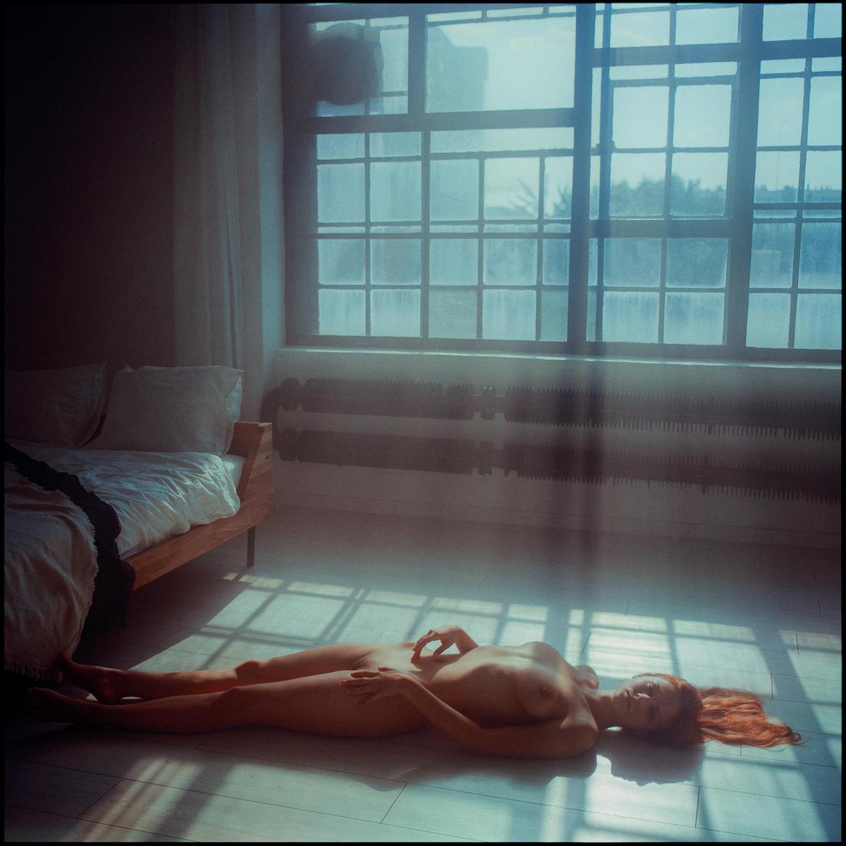 A nude woman with red hair lies on the floor in a room with a large window and a bed.