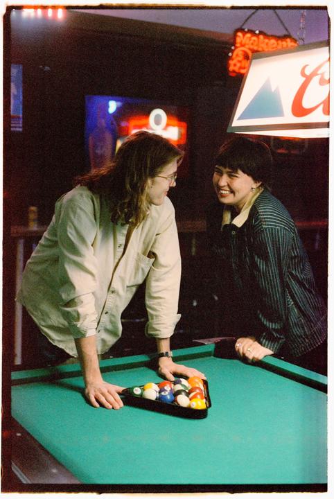 Two women are smiling at each other while leaning over a pool table with racked balls in a dimly lit bar.