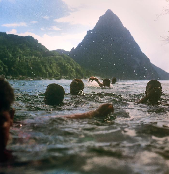 Several people swim in the ocean with a large mountain in the background.