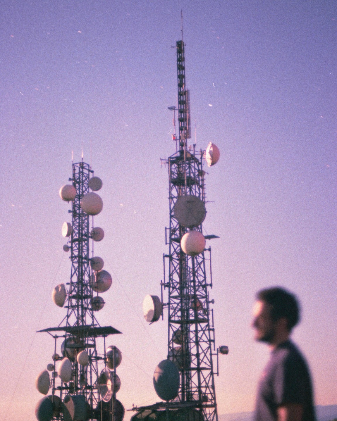 A man stands in front of two large communication towers against a purple sky.