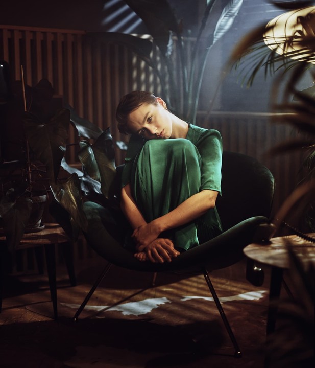 A woman in a green dress sits curled up in a chair surrounded by plants, with dramatic shadows cast across the room.