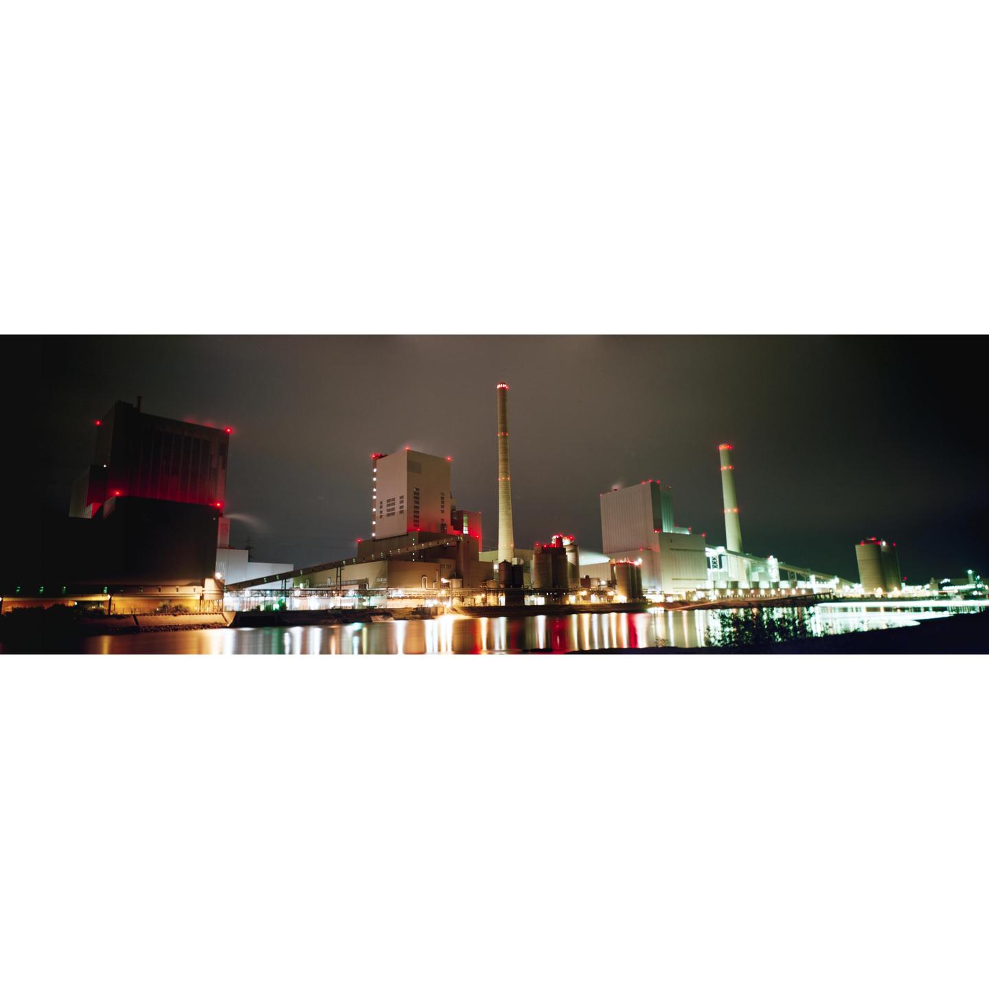 A power plant with multiple smokestacks and buildings is illuminated at night, reflecting its lights on the water.