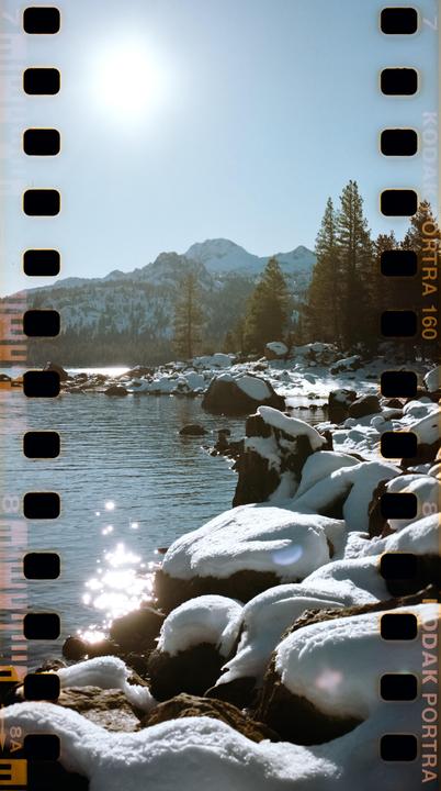 A bright sun shines over a snowy mountain lake with pine trees and snow-covered rocks along the shore.
