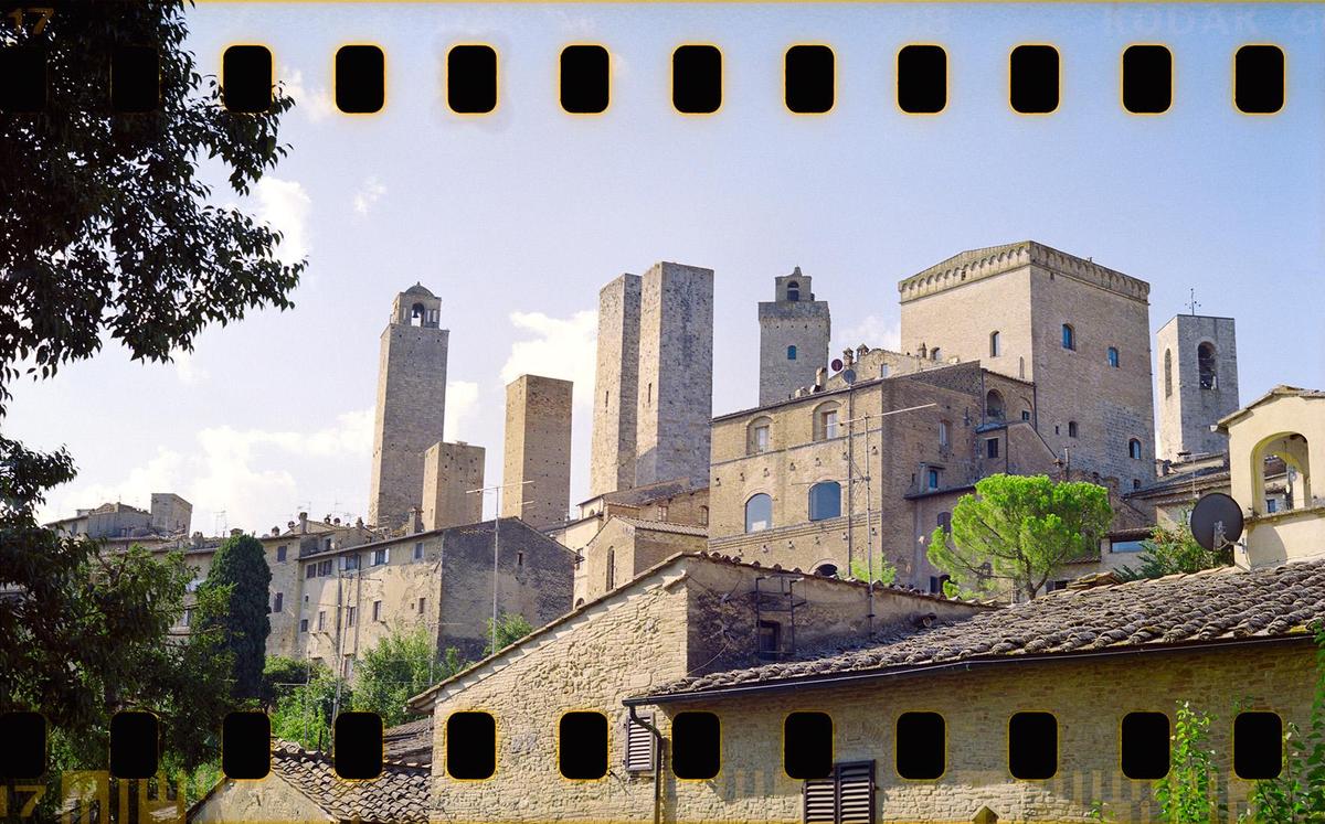 A medieval town with multiple stone towers rises above a hillside with trees and houses.