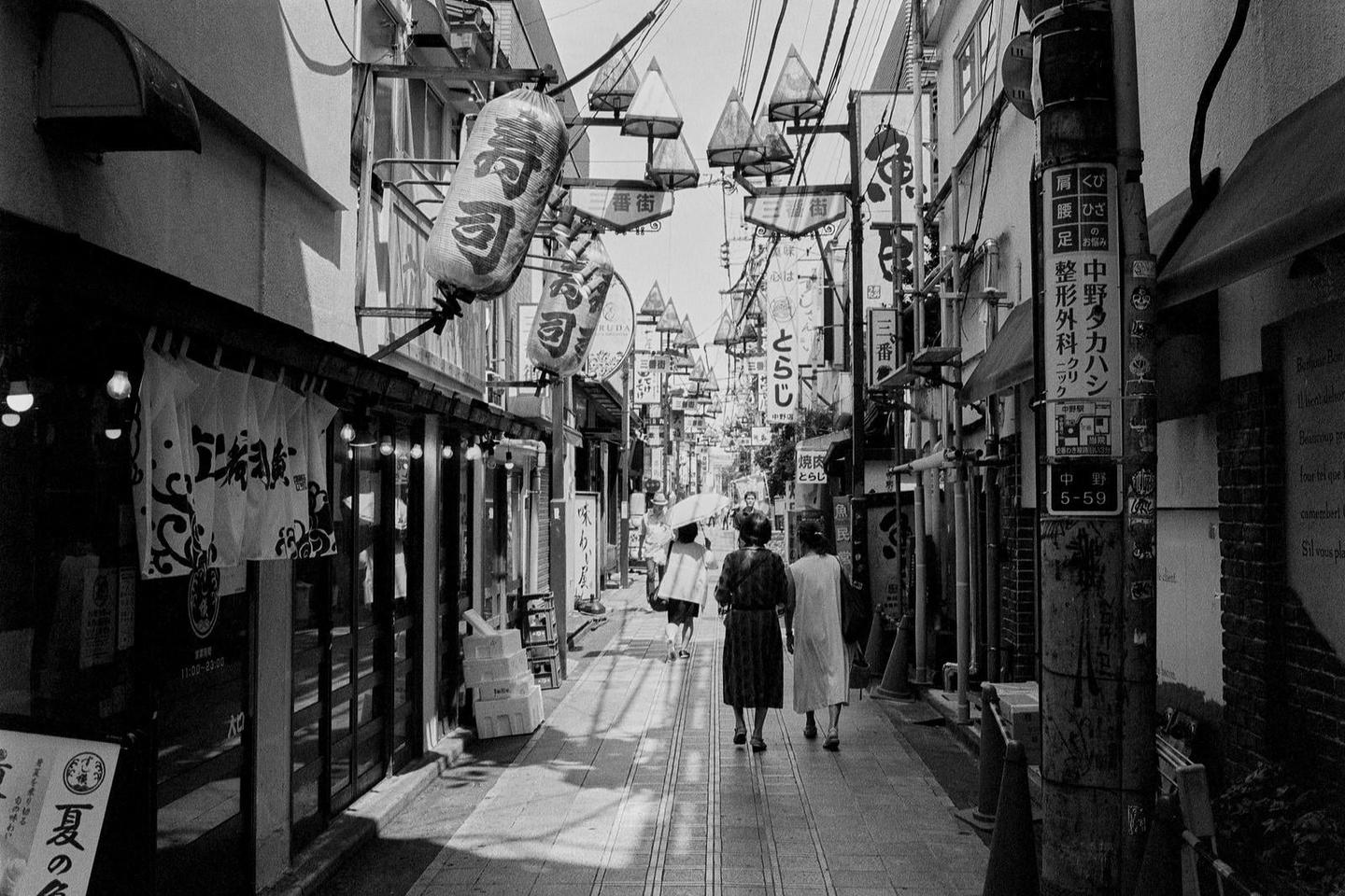 Three women walk away from the viewer down a narrow street lined with shops and signs.