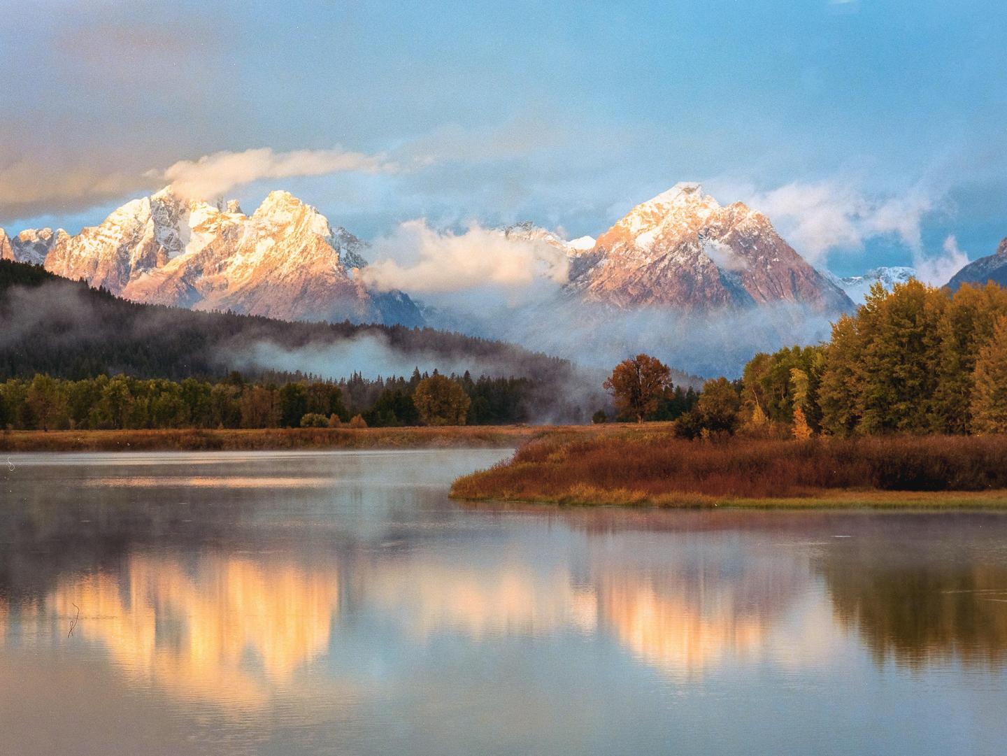 Snow-capped mountains rise above a river and forest, reflecting in the water under a cloudy sky.