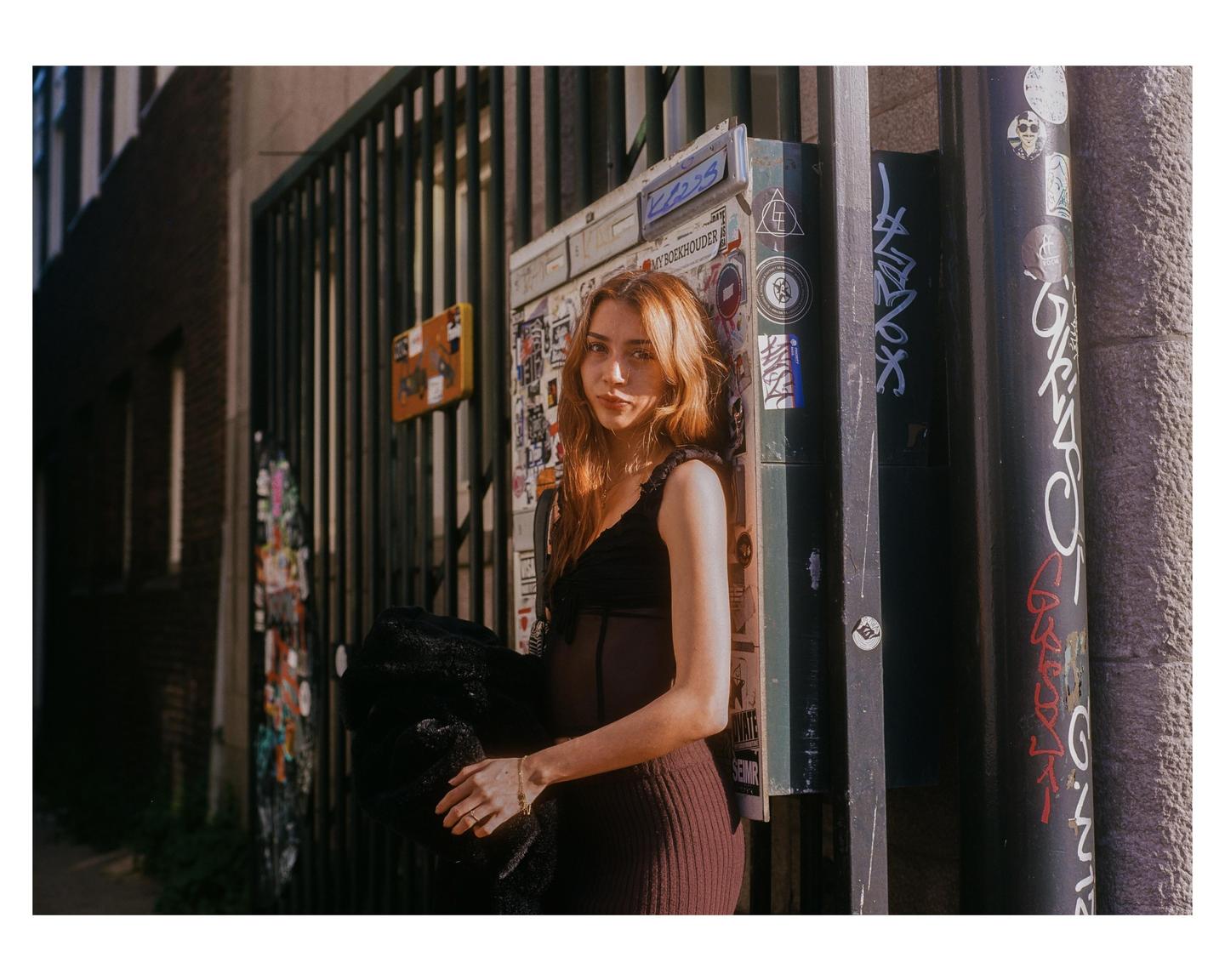 A woman with long hair stands next to a metal gate and a utility box covered in stickers and graffiti.