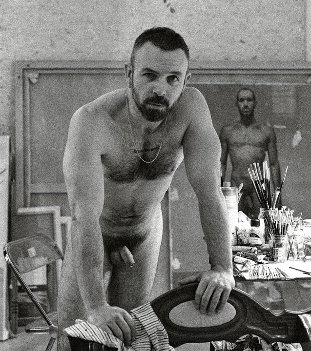 A nude man with a beard leans on a chair in an art studio, with a painting of another nude man in the background.