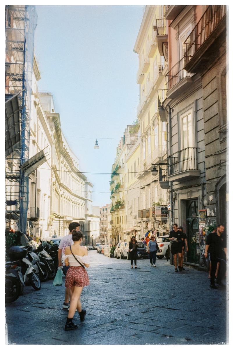 A narrow street lined with tall buildings and parked scooters is filled with people walking under a clear sky.