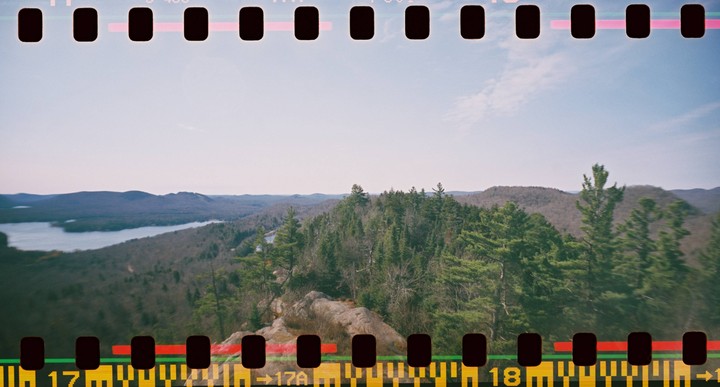A panoramic landscape shows a lake surrounded by hills and evergreen trees under a clear sky.