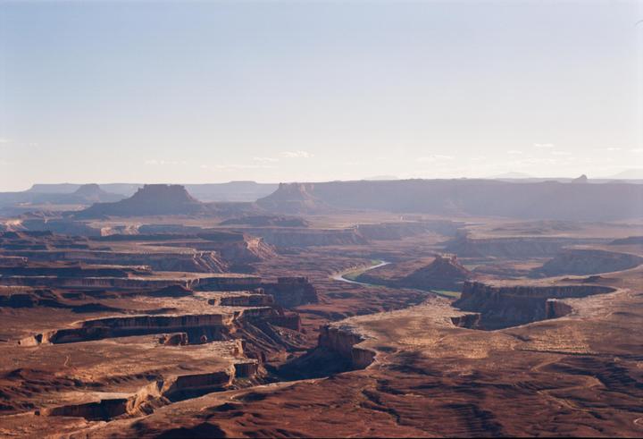 A wide landscape shows a winding river through a vast canyon under a clear sky.